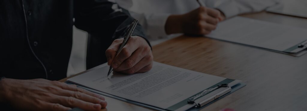 Two people, possibly consumer protection attorneys, review and write on documents using pens and clipboards at a desk.