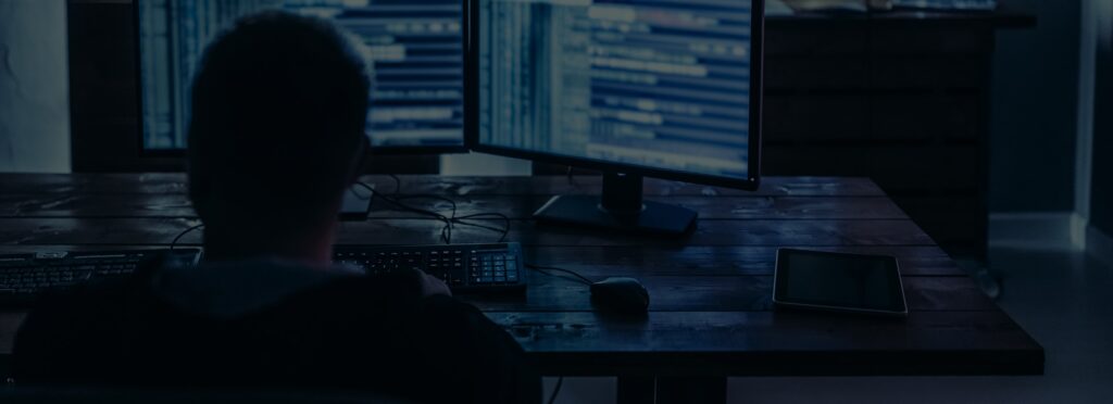 A dedicated person sits at a desk, coding on dual monitors in a dim room, focused with precision like identity theft lawyers.